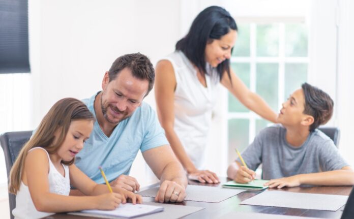 Padre y madre acompañan a sus dos hijos mientras realizan tareas escolares alrededor de la mesa del comedor, reflejando la importancia del ejemplo y la convivencia familiar.