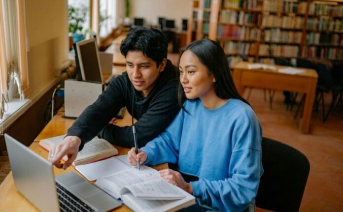 Dos estudiantes universitarios estudian en una biblioteca utilizando libros y una computadora portátil como apoyo para su aprendizaje.
